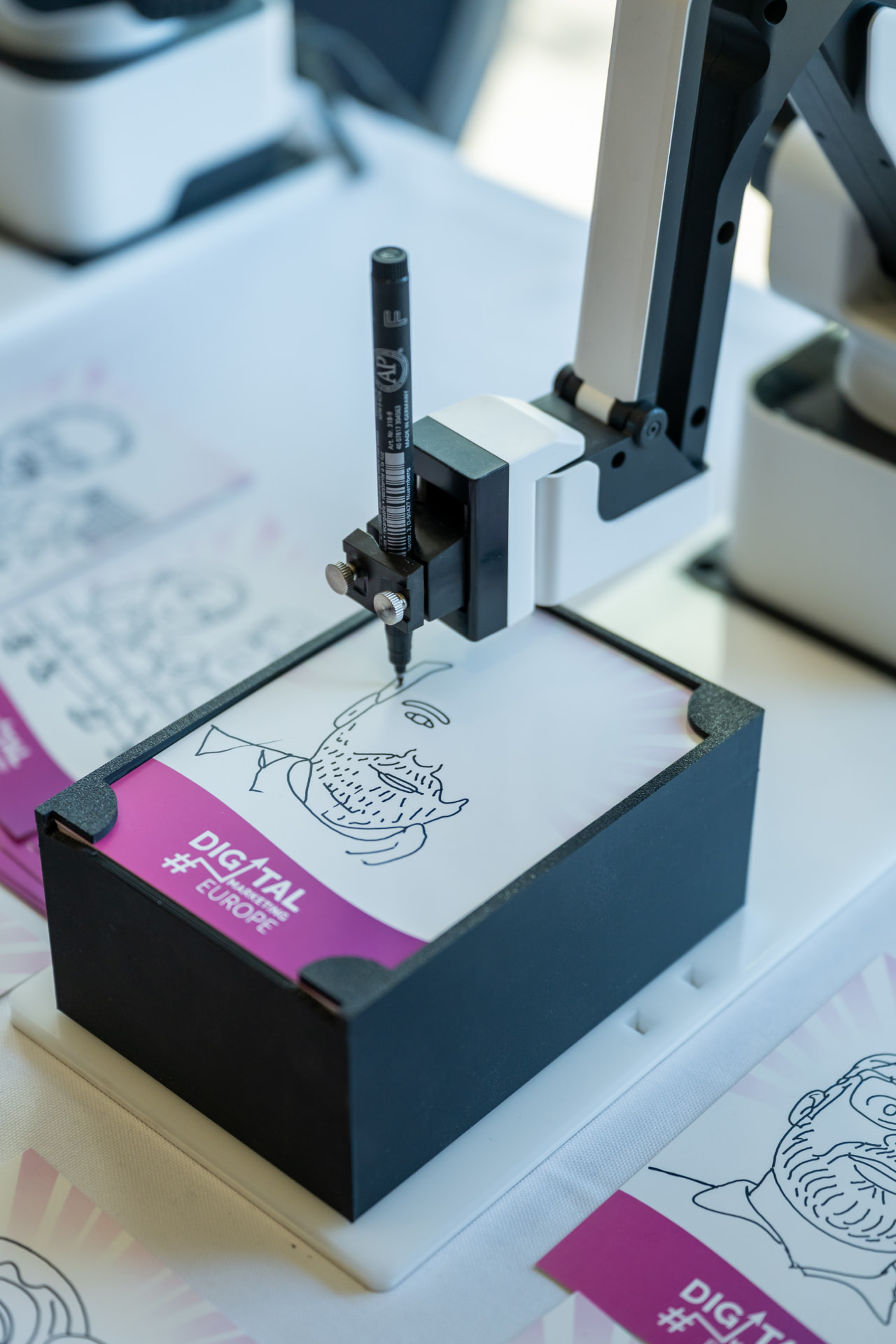 Robotic arm drawing a portrait with a marker on a branded event card