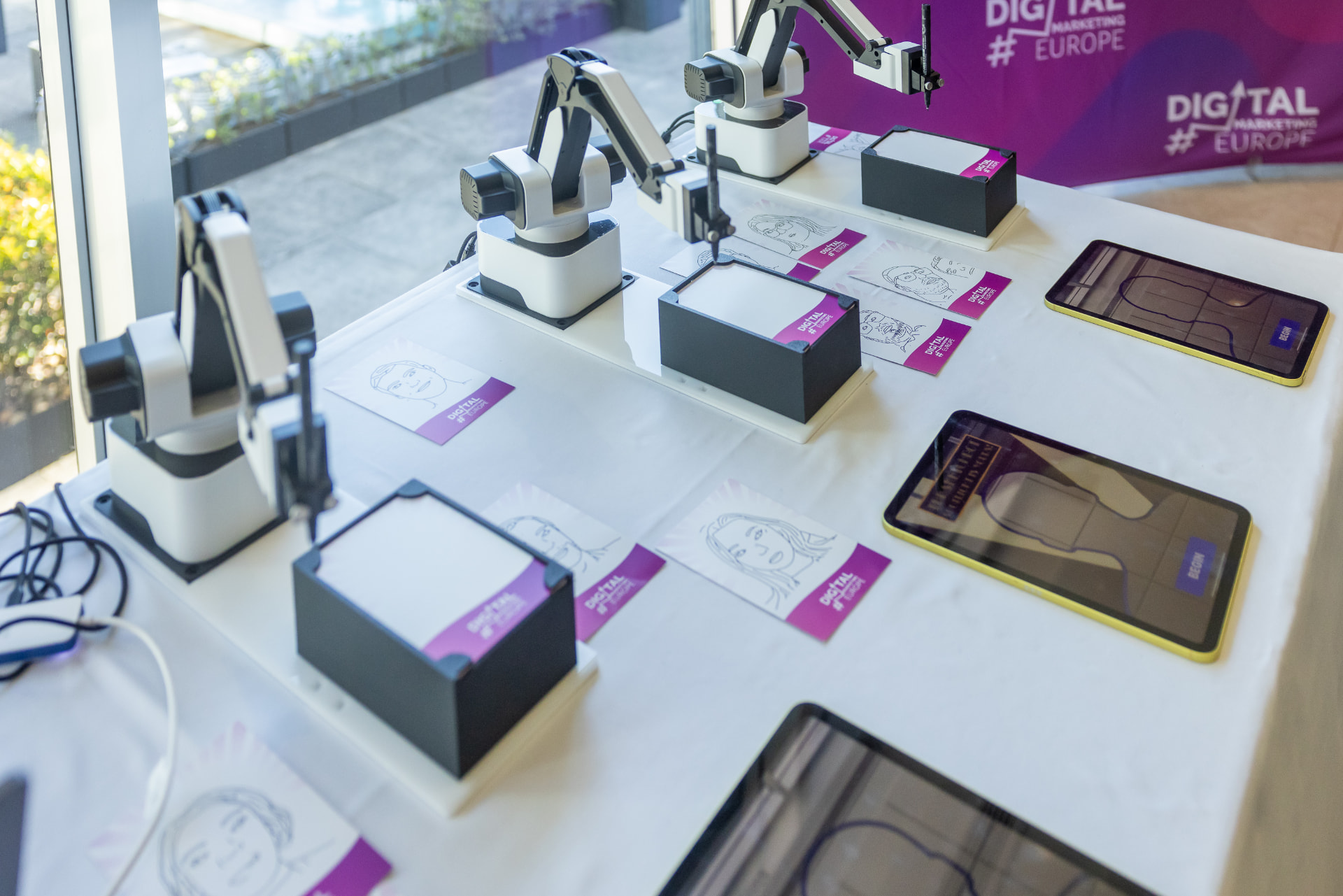 Three robotic arms set up side by side on an event table with drawn portraits
